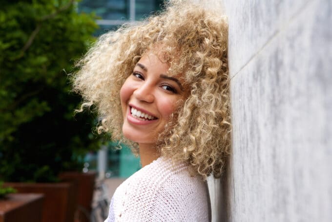 Black women with type 3B curly hair dyed blonde with semi-permanent hair dye leaning on a stone wall.
