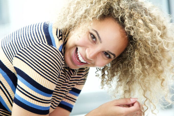 American women with ombre waves styled with permanent dye wearing a black, yellow, and blue fitted short-sleeve sweater with a huge smile.