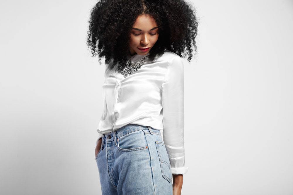 American female wearing a white silk shirt, silver necklace, faded blue jeans, and red lipstick with wavy natural hair.