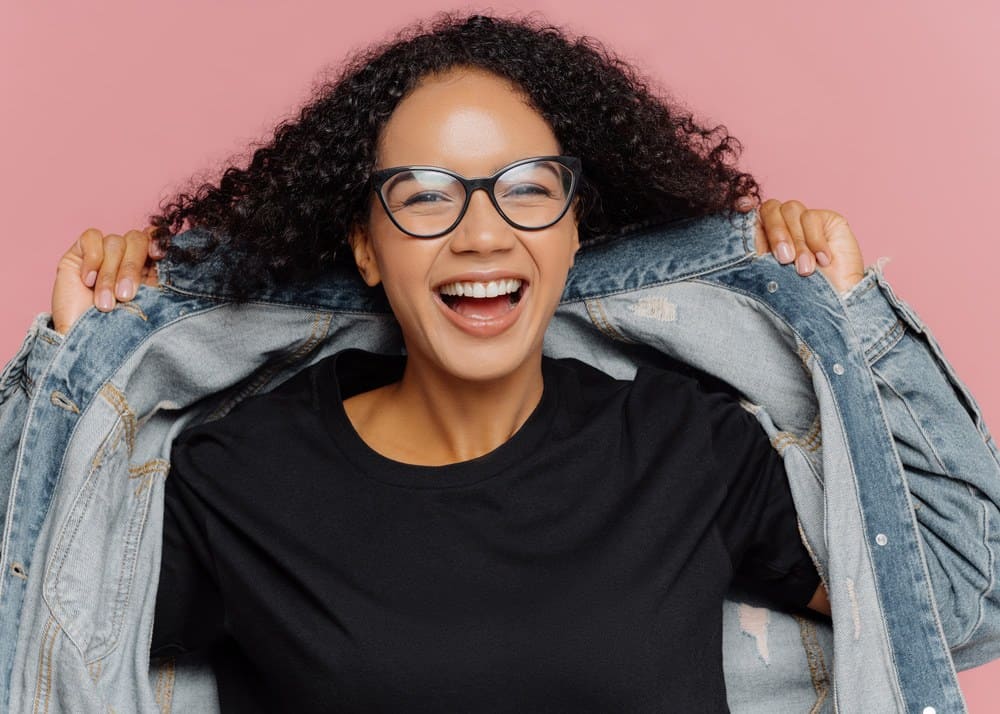 Back lady with a huge smile wearing a blue jean jacket and type 3B curly hair with a black undershirt.