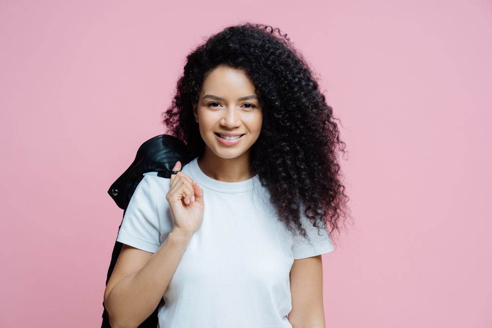 Ethnic African American woman with curly hair wearing a white t-shirt and carrying a black leather jacket over her shoulder.