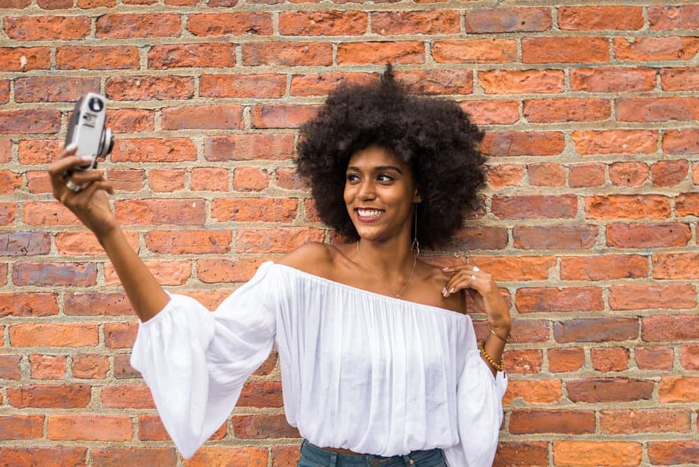 Black female wearing a white crop top with type 4C hair taking a selfie with a camera against a bricked wall.