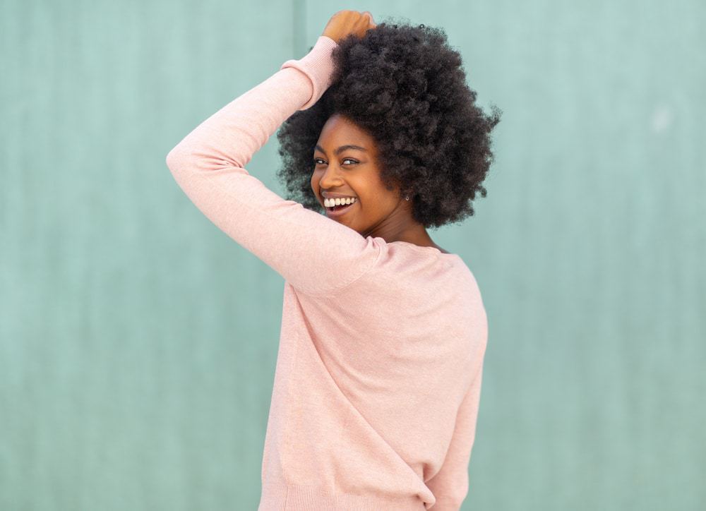 African American women with naturally curly hair looking back at the camera with a huge smile, wearing a oink sweater and earrings.