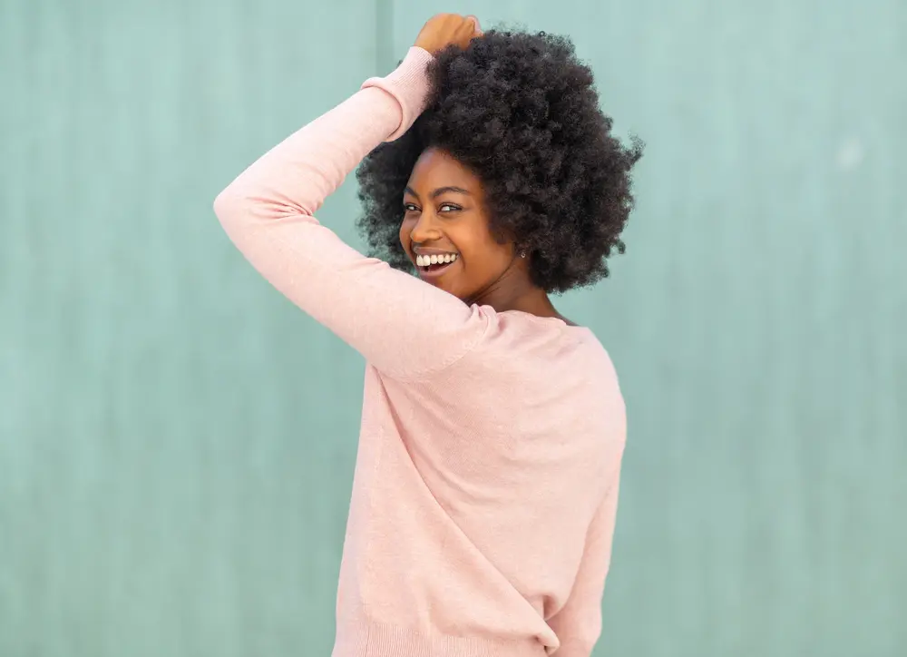 African American women with naturally curly hair looking back at the camera with a huge smile, wearing a oink sweater and earrings.