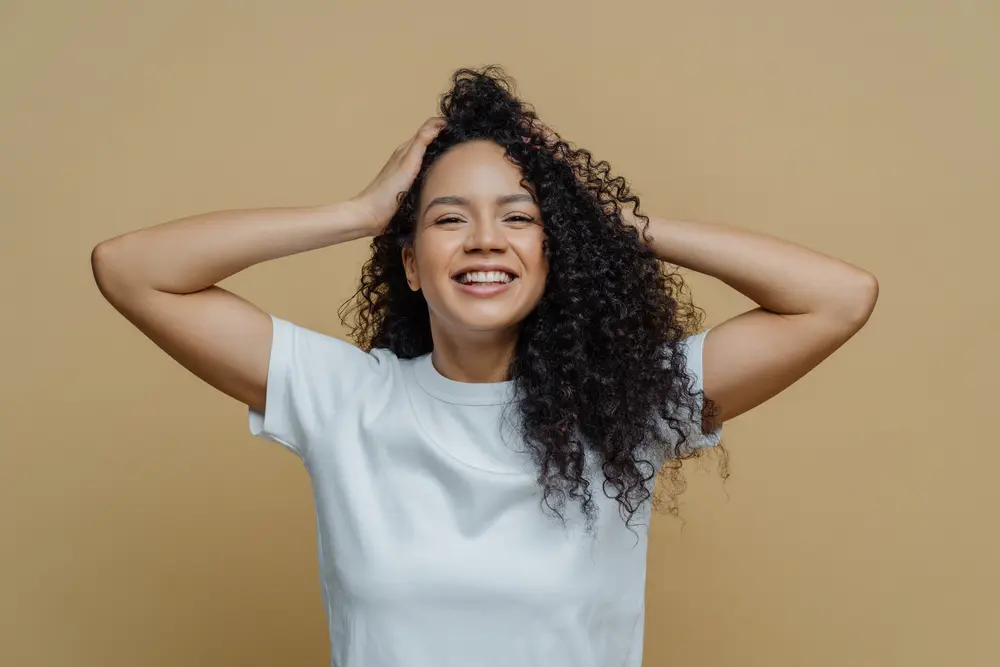 Happy black girl with curly hair holding her hair strands that were moisturized with bergamot oil while wearing a white t-shirt.