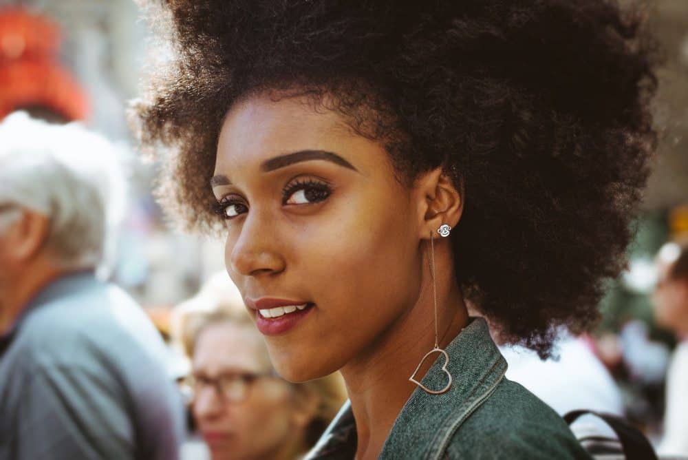 African American female standing in a crowd wearing one heart-shaped earring and one bear-shaped earring with red lipstick and a blue jean jacket. 