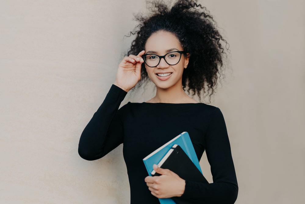 Cute black female with frizzy hair wearing a black sweater, reading glasses and carrying two hair care journals.