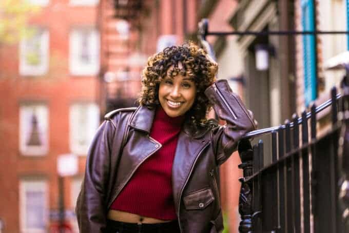 Adult female with ombre curls wearing a brown leather jacket and red ribbed sweater