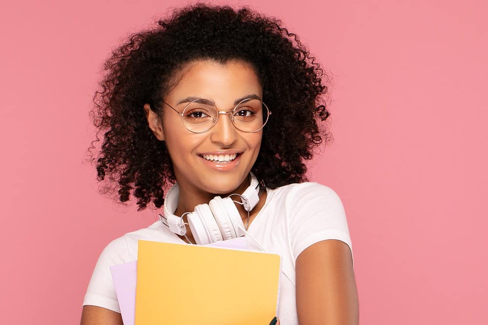 Cute black woman with round eye glasses, pink lipstick, curly, white Dr. Dre headphones, and a yellow notebook.