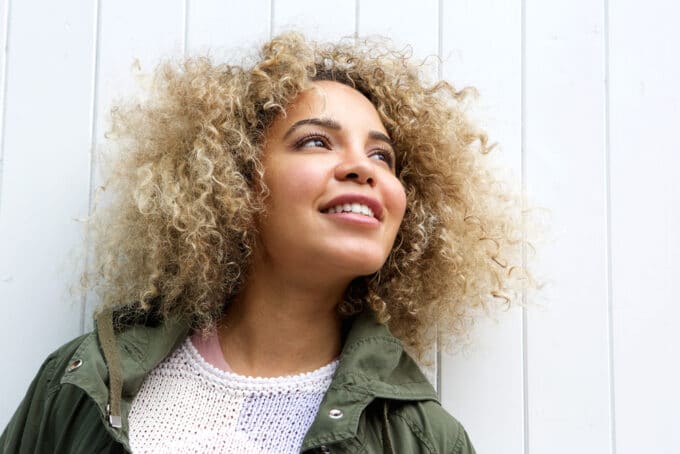 Beautiful black female standing in direct sunlight with blonde type 3b curly hair wearing a green jacket and white sweater.