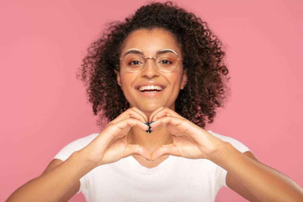 Female wearing a white t-shirt, eye shadow, and black fingernail polish, making a heart symbol with her hands.