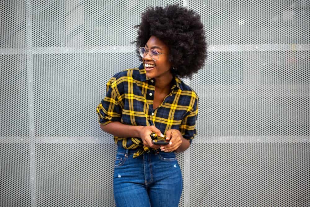 Black girl wearing blue jeans with a blue and yellow shirt and glasses while using a mobile phone.