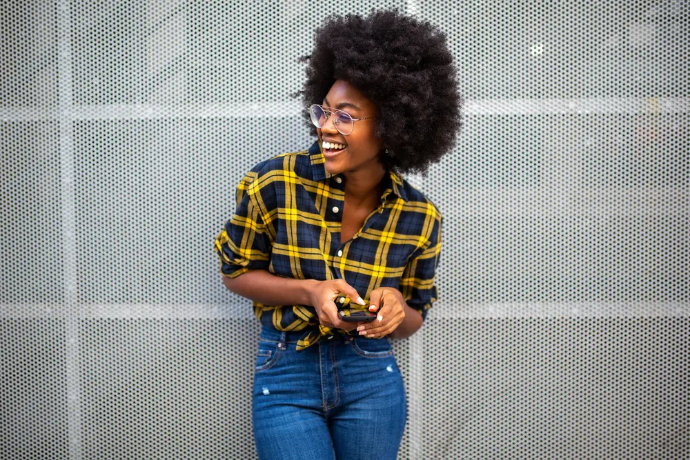 How To Grow Natural Hair Faster: Grow Hair Fast Step-by-Step 6 Black girl wearing blue jeans with a blue and yellow shirt and glasses while using a mobile phone.