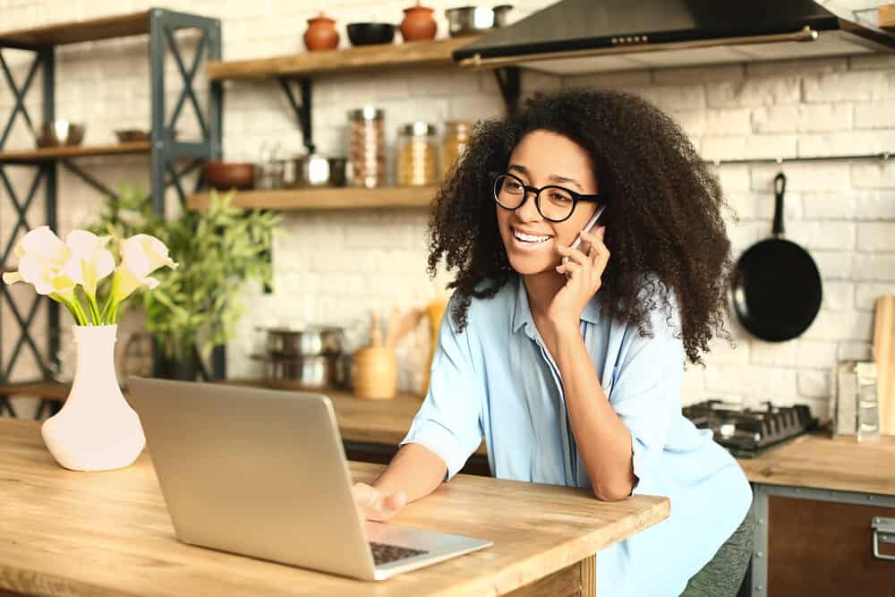 A woman in glasses and a blue shirt stands at a kitchen counter on the phone, one hand on an open laptop