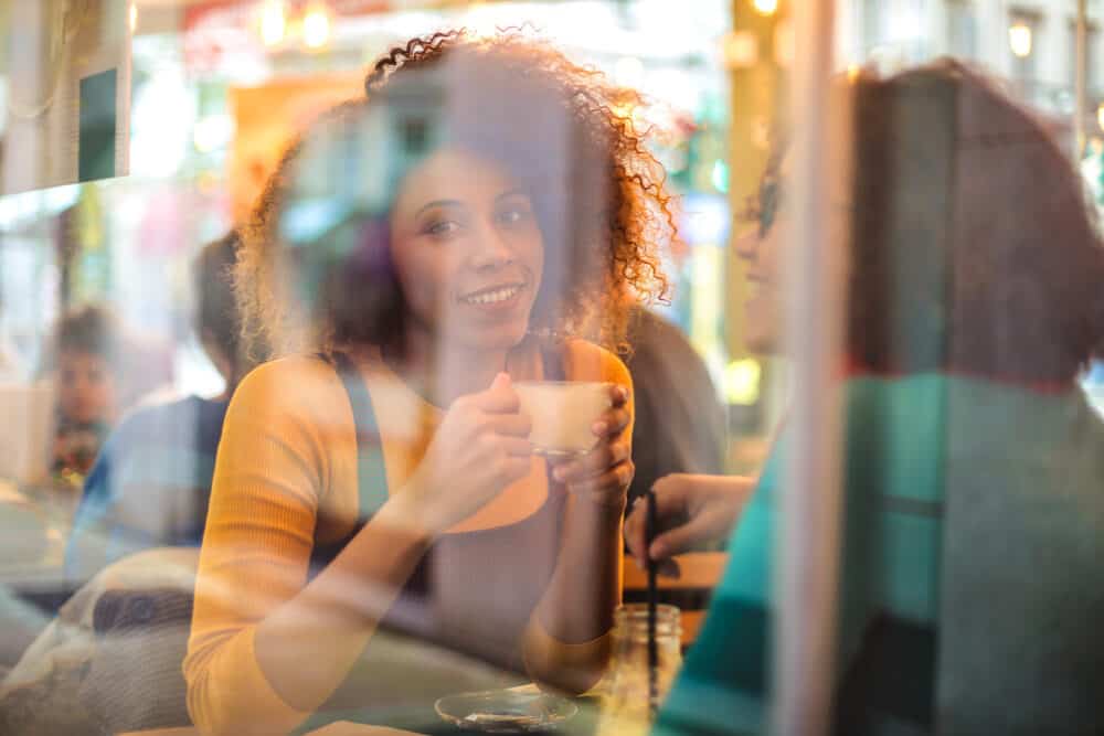Two women at a cafe table seen through a window, one with curly hair holding a cup and smiling
