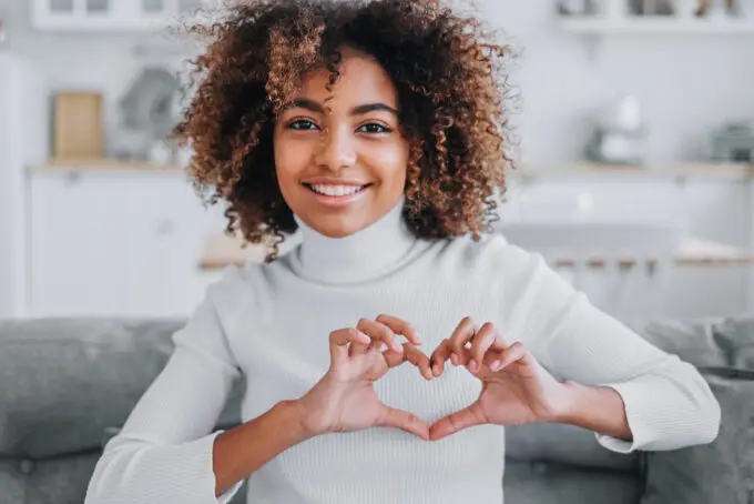 Black female with type 3c curly hair smiling while making a heart symbol with her hands. 