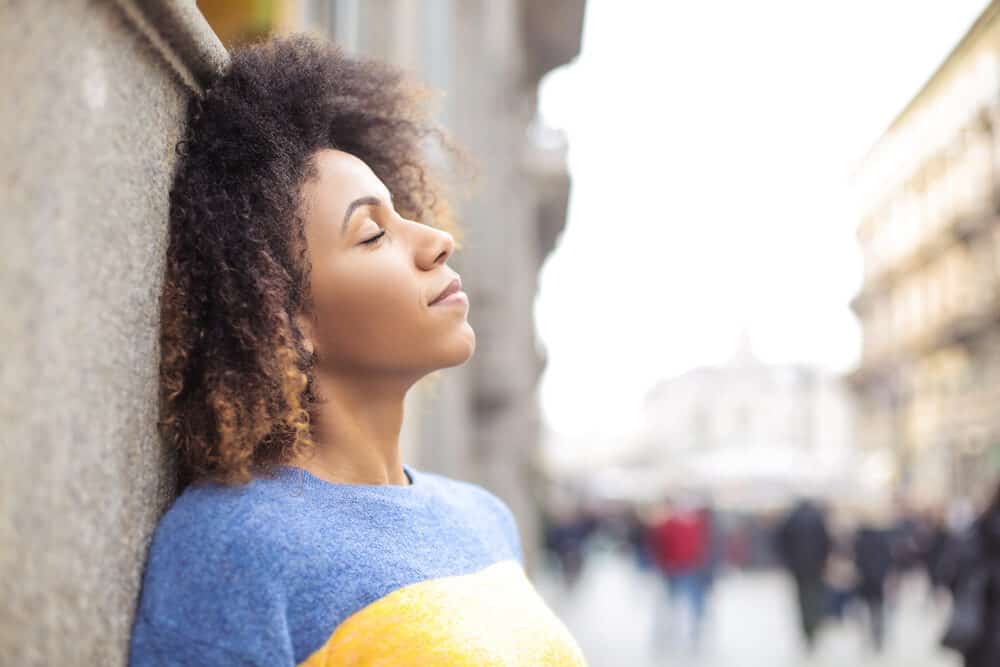 Woman with curly hair leaning against a stone wall with her eyes closed, in a blue and yellow sweater