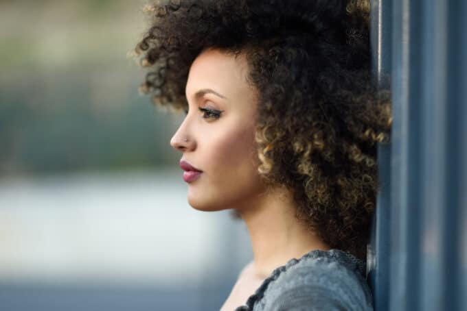 American female with type 4B curly hair, a small nose ring, red lipstick while leaning up against the wall.