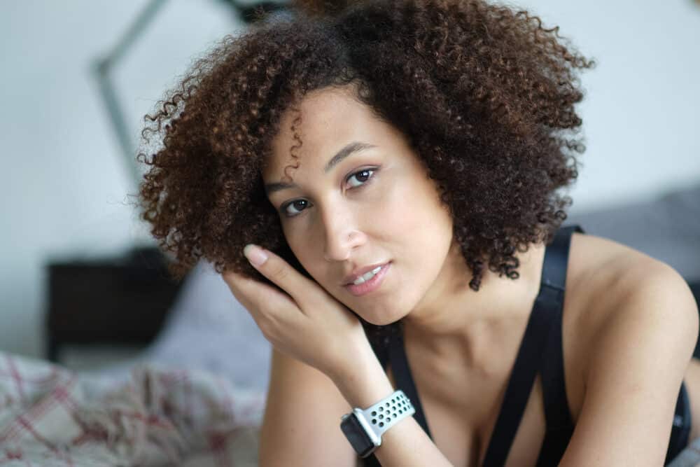 A woman with a brown curly afro resting her head on her hand on a bed, in a black strappy top and white watch