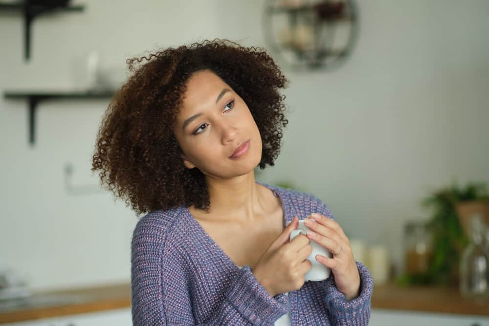 A woman holding a white mug with both hands in a kitchen, wearing a purple knit cardigan