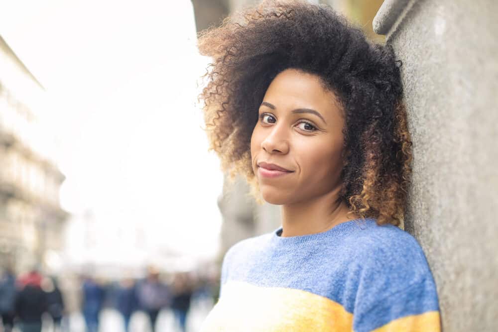 Woman with a curly afro leaning against a stone column on a city street in a blue and yellow sweater