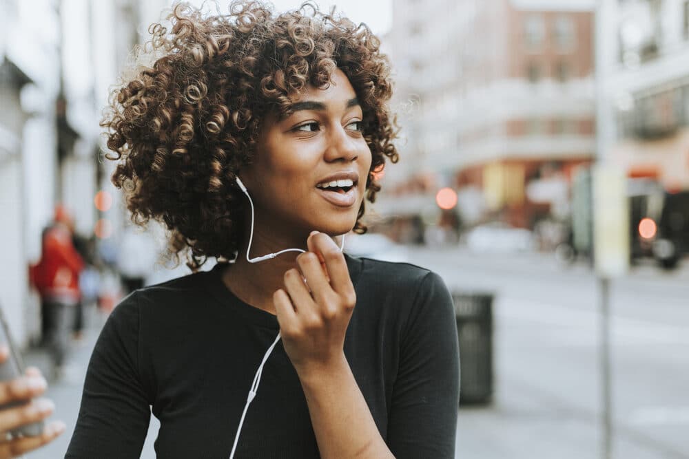 Beautiful American American female wearing a black shirt with naturally curly hair strands using earphones during a phone call.