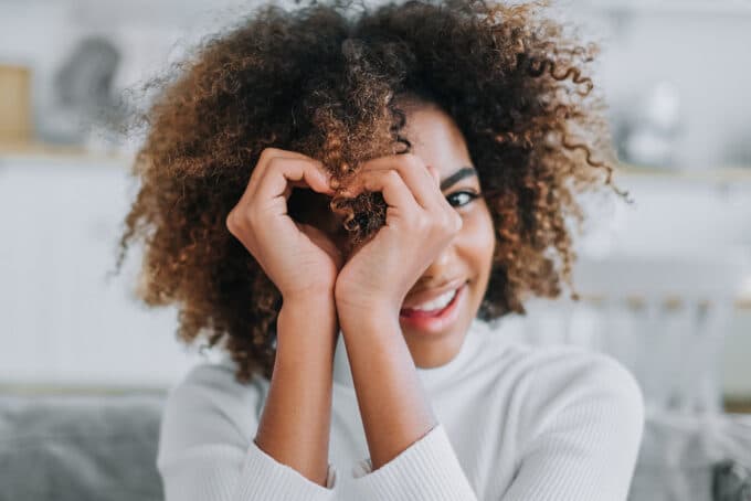 Light-skinned black female making a heart symbol with her hands, with naturally curly hair.