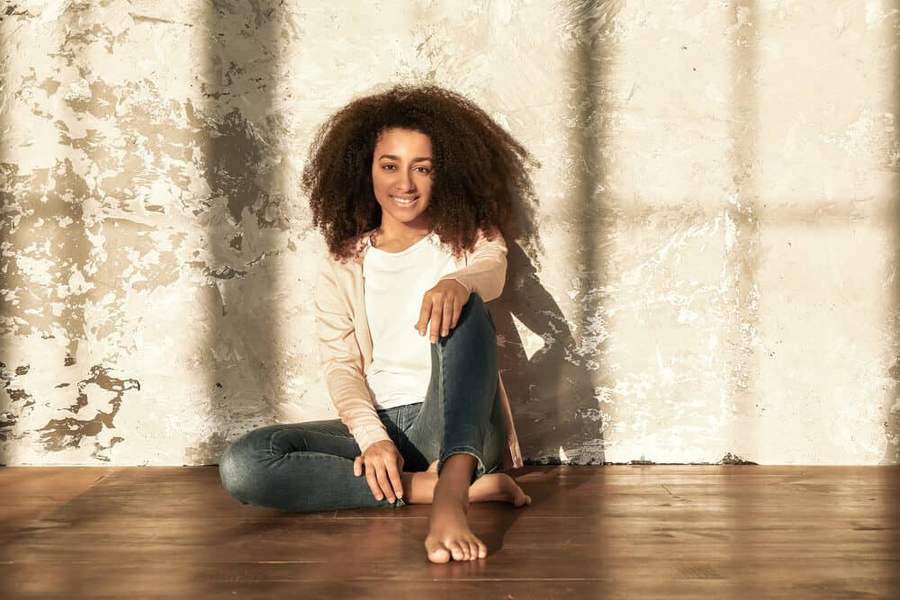 A woman sits cross-legged and barefoot on a wood floor against a pale textured wall, curls framing her face