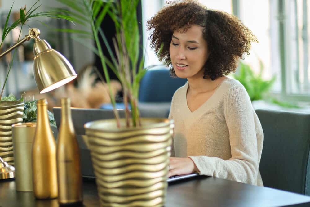 A woman with a curly afro typing at a dark desk beside a gold planter and a brass lamp, in a cream sweater