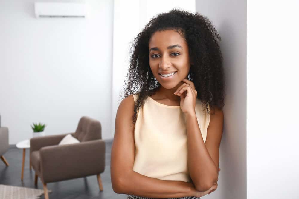A woman leans against a white wall with her hand near her chin, long curls over one shoulder, in a cream top