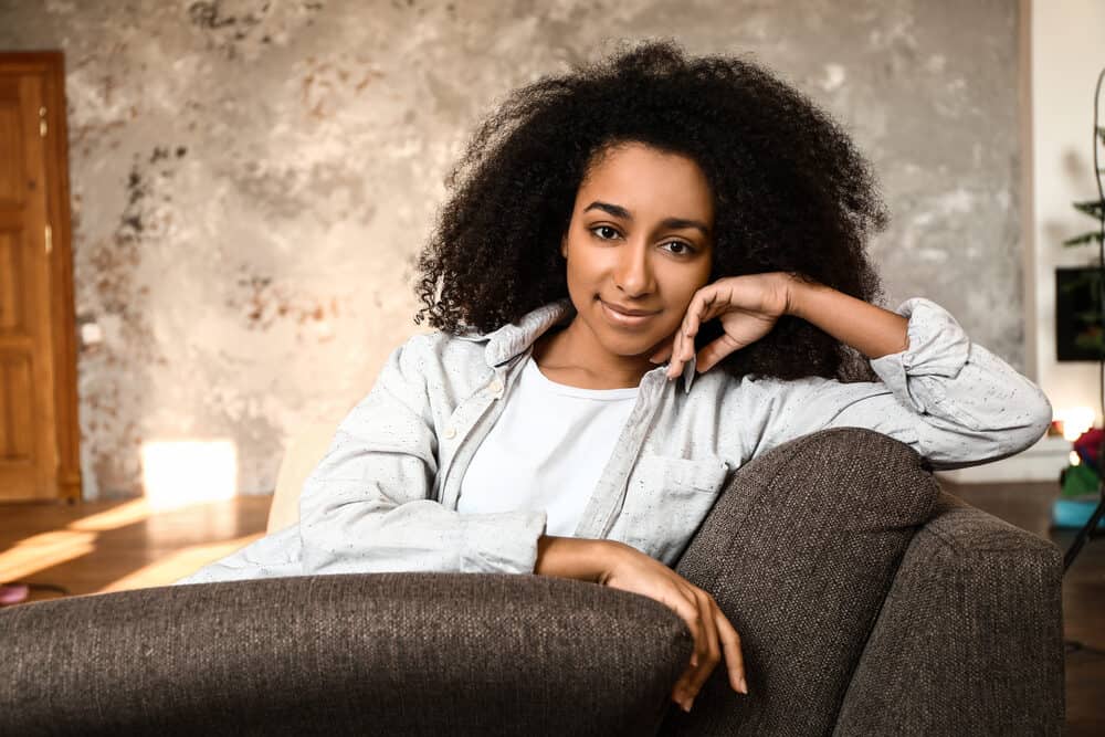 A woman with a curly afro leans on the arm of a couch with her chin on her hand, in a speckled shirt over a white tee
