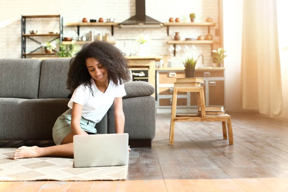 A woman with a short curly afro sits on the floor beside a couch using a laptop, in a white tee and khaki skirt