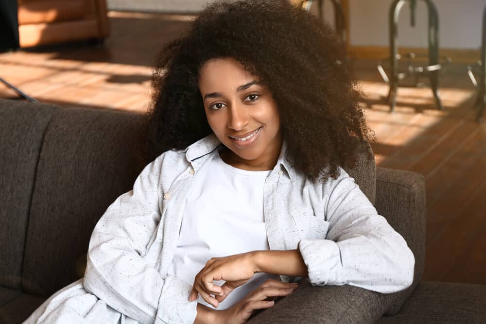 A woman sits back on the couch smiling, curls loose around her shoulders, hands resting in her lap