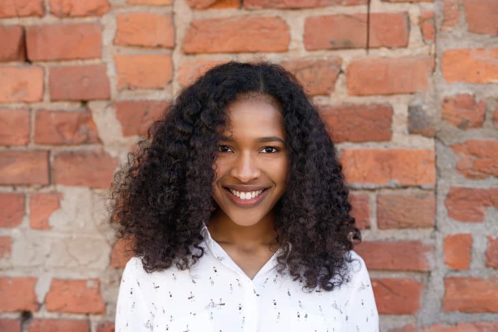 Black woman with a big smile standing in front of a bricked wall with very curly hair strands.