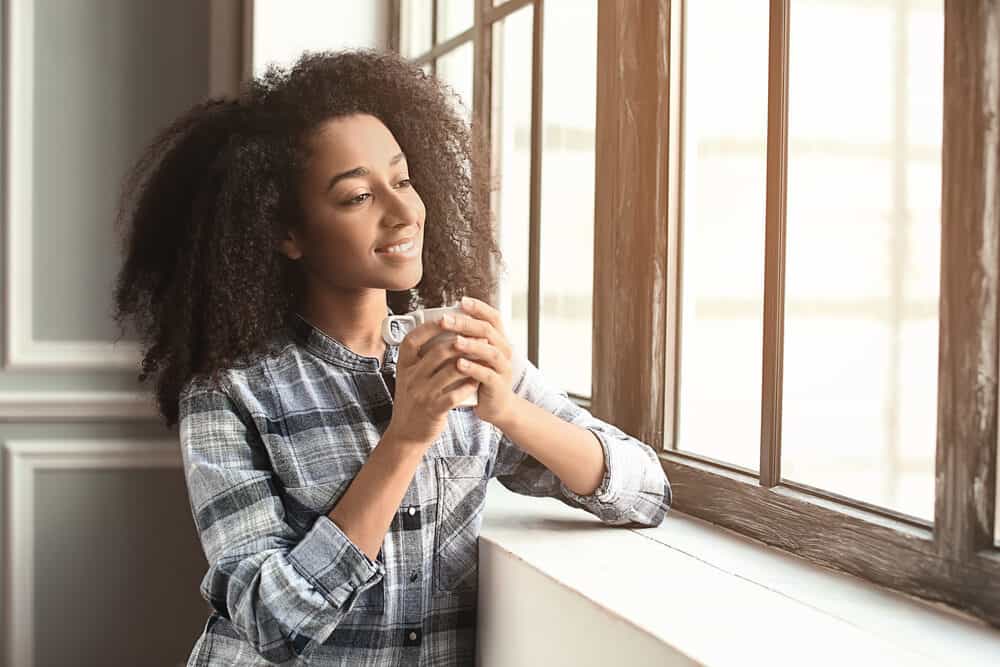 A woman with curly hair holds a mug in both hands beside a window, wearing a plaid shirt and looking outside