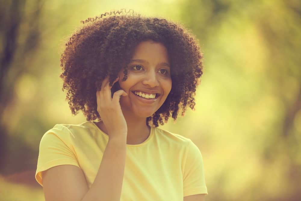 Black female with a yellow shirt talking on her new iPhone outdoors.