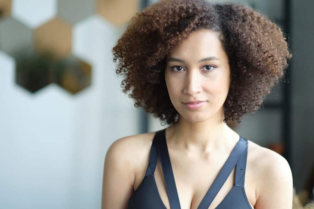 A woman in a gray strappy top in front of a white hexagon tiled wall, curls framing her face