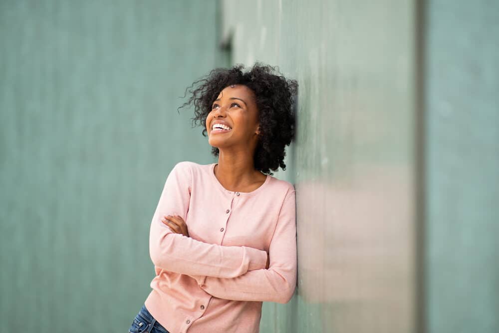 African American female leaning against a green marble wall wearing a pink shirt and casual pants.