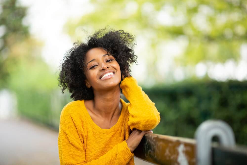 Female standing outside leaning against a rusty iron fence wearing a yellow shirt with naturally curly hair.