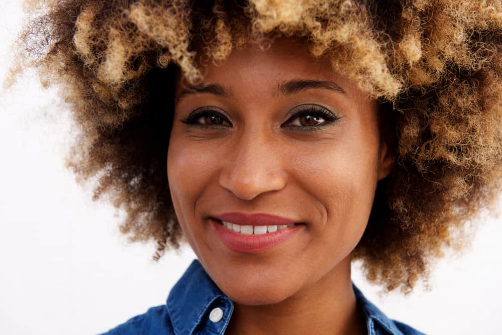 Black women with ombre curly hair wearing a blue collared shirt, eye shadow, and red lipstick.