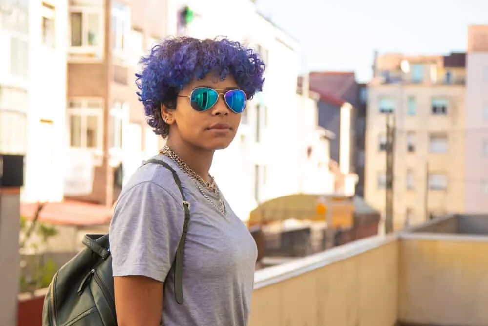 How to Dye Dark Hair Purple With and Without Bleach at Home 8 Latin female standing outside wearing a gray t-shirt, neck chains, and a green backpack.