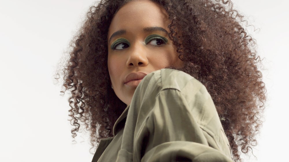 African American female with medium to long hair styled with sea salt texturizing spray wearing metallic green eyeshadow looking back over her shoulder.