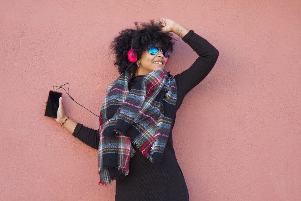 Black women dancing in the street while wearing a black dress, headphones, and a multi-colored winter scarf.