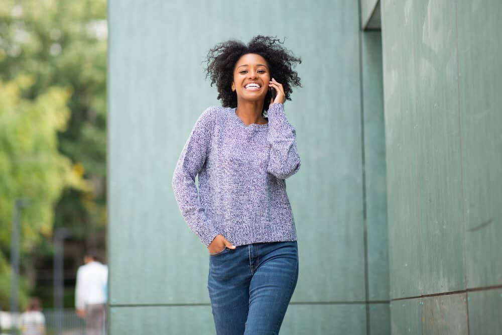 Women walking outside with curly hair talking on the phone wearing a casual sweater and blue jeans.