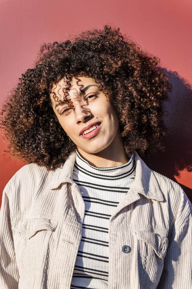 Latina women with a type 3 curly hairstyle wearing a corduroy jacket, white and black striped undershirt, and pink lipstick.