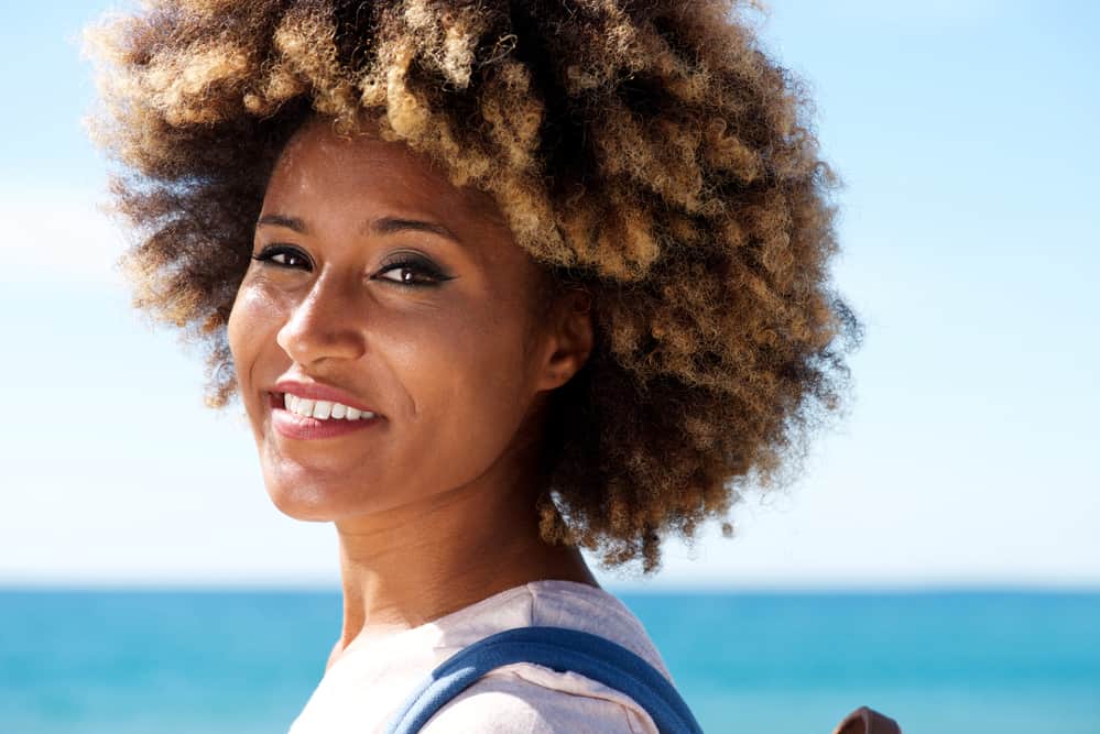 Black female standing in front of the beach with blonde naturally curly hair strands wearing a pink t-shirt and blue backpack.