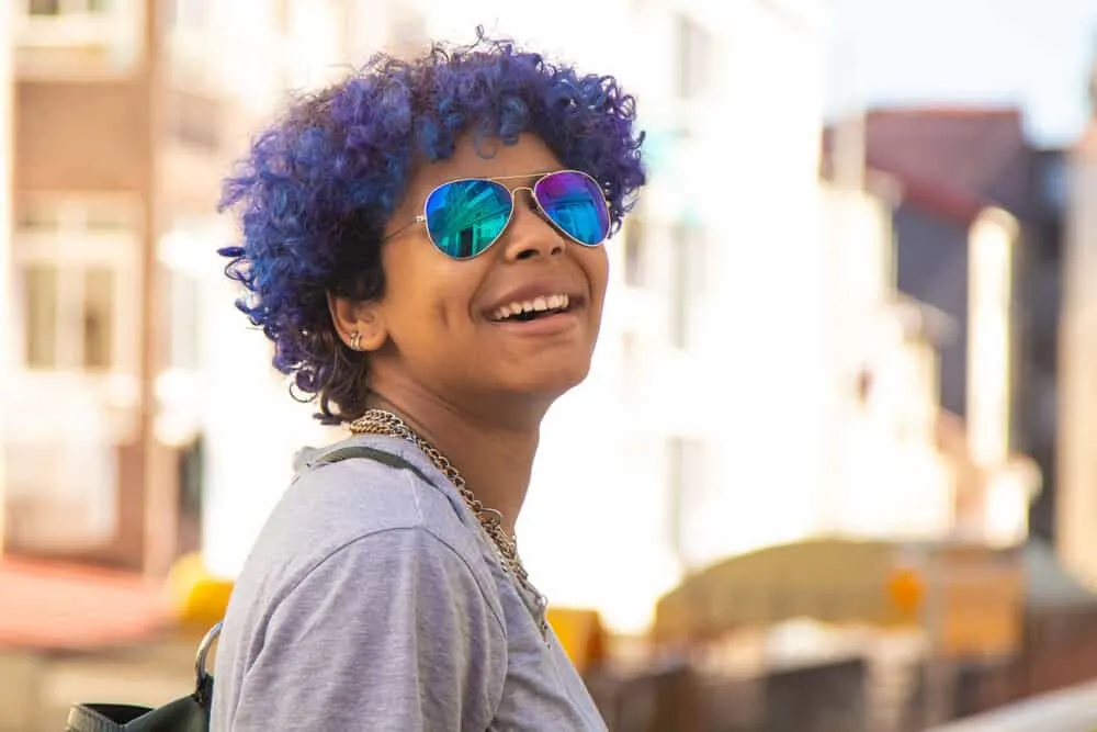 How to Dye Dark Hair Purple With and Without Bleach at Home 4 Hispanic female with blue hair looking back over her shoulder and smiling at the camera.