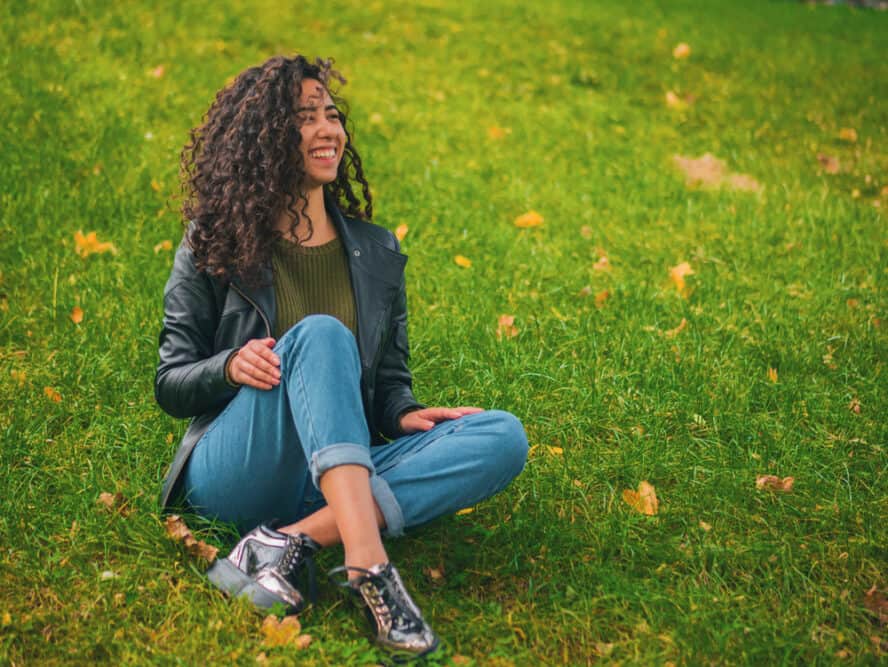 Beautiful brunette woman with very long curly hair sitting outside in the grass with her legs crossed.