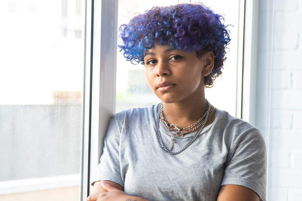 Latin girl leaning against a window seal wearing multiple necklaces and a gray t-shirt.