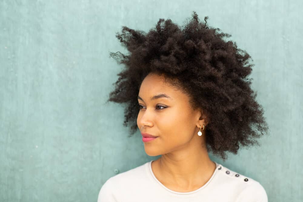 Female with a naturally curly hair type standing against a green wall wearing red lipstick and pearl earrings.